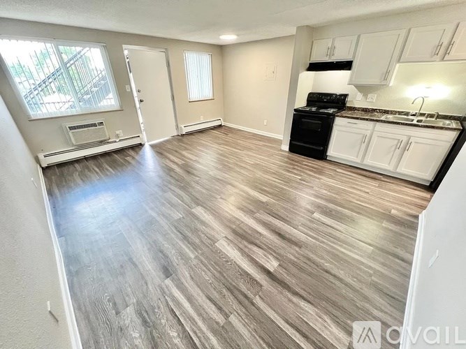 A spacious living room with wood flooring and a kitchen area with white cabinets and black appliances.