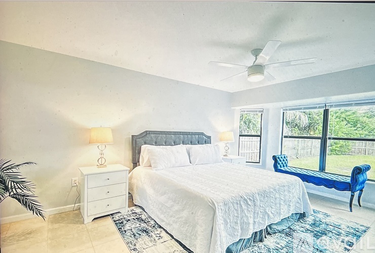 A bedroom with a large bed, a ceiling fan, and a view of the backyard through the window.