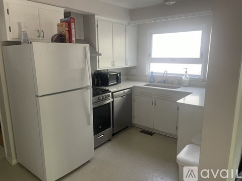 A white refrigerator stands in a kitchen with white cabinets and a window.