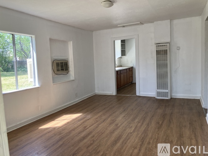 A room with wooden flooring and white walls, featuring a window, a mirror, and a wooden cabinet.