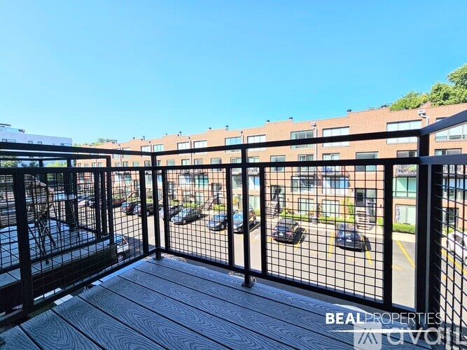 A balcony with a black railing and wooden flooring overlooks a parking lot and a building.