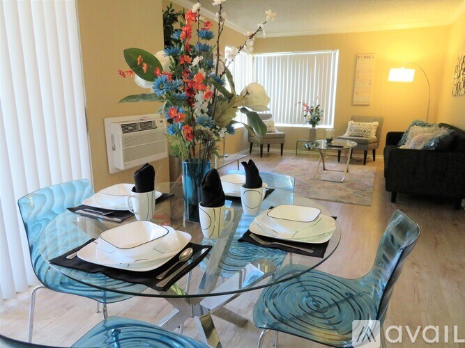 A dining table set with plates and glasses is in the foreground of a bright room with a sofa and chairs in the background.