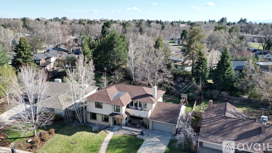 A large house with a brown roof is surrounded by trees and other houses in the distance.