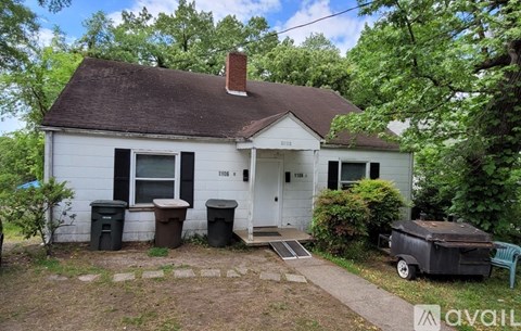 A white house with a brown roof and a chimney is surrounded by trees and has a trash can and a grill in front of it.
