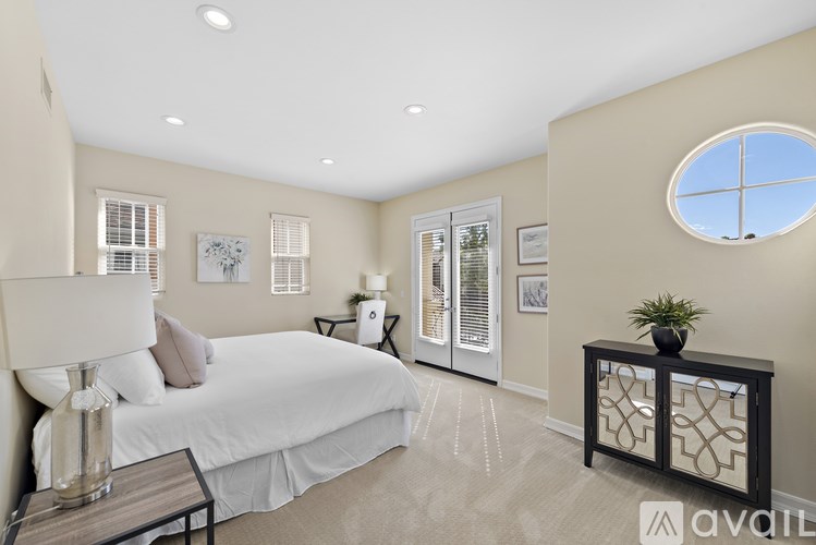 A bedroom with a large bed, a nightstand with a lamp, a window with blinds, a round window, a plant on a table, and a framed picture on the wall.