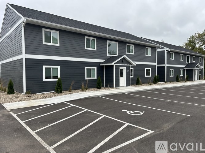 A parking lot in front of a grey building with trees in the background.