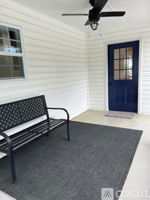 A black bench with a patterned back sits on a grey rug in front of a white house with a blue door.