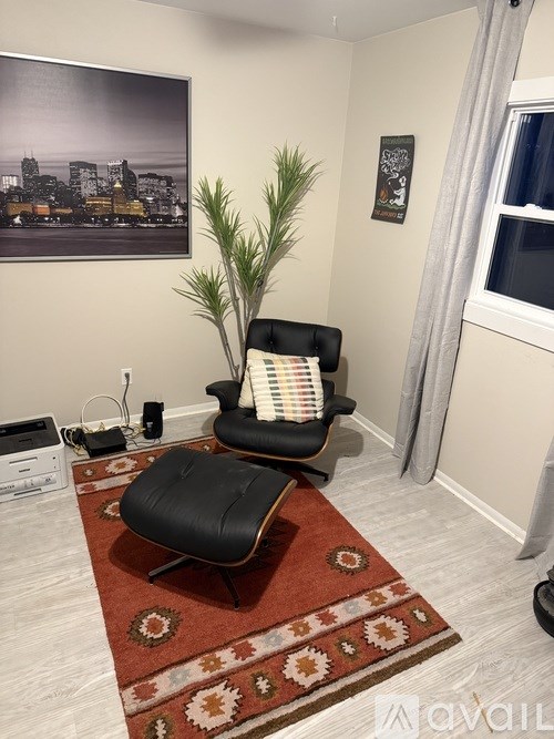 A black chair with a striped pillow sits on a red and beige rug in a room with a cityscape poster on the wall.