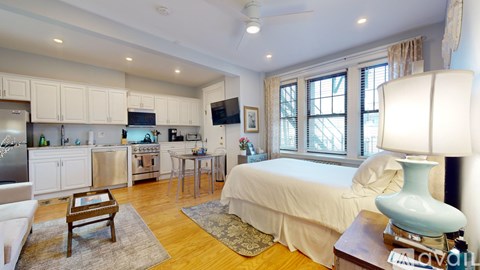 A well-lit bedroom with a bed, a table with a lamp, and a kitchen area with appliances.