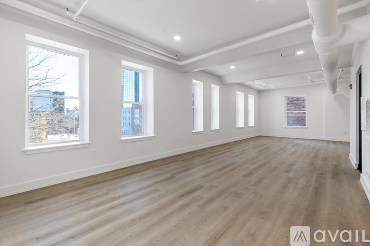 A spacious room with wooden flooring and white walls, featuring a large window with a view of a cityscape.