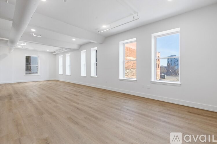 A spacious room with wooden flooring and white walls, with three windows and a view of buildings outside.