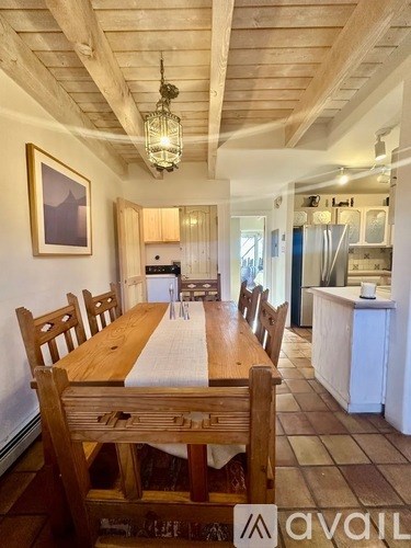 A wooden dining table with chairs is set up in a room with a kitchen in the background.