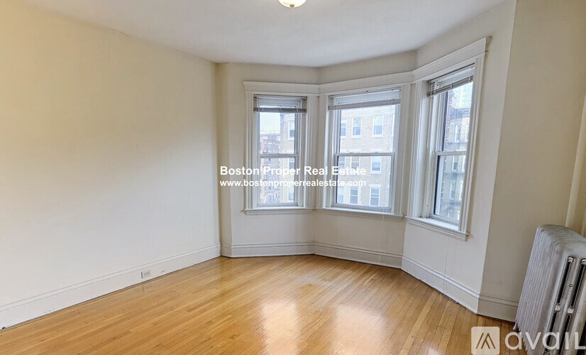 A room with wooden floors and white walls, featuring a window with a view of buildings outside.