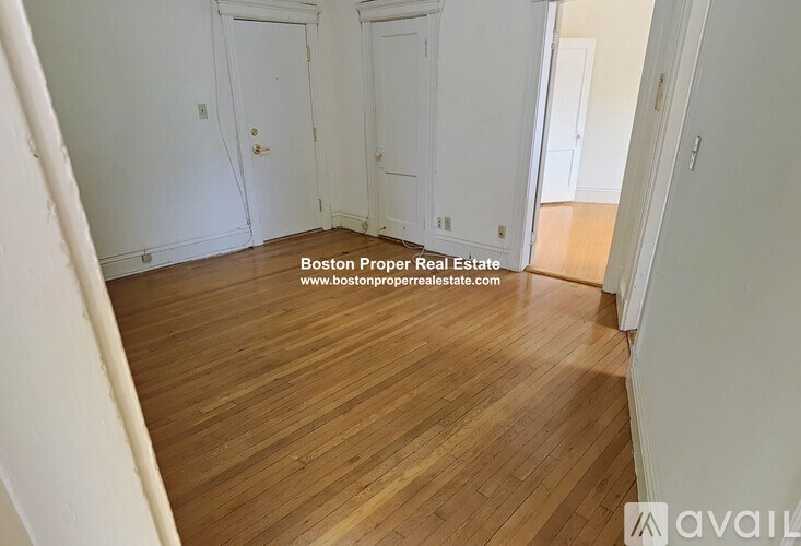 A room with wooden flooring and white walls, with a door on the left and a partially visible door on the right.