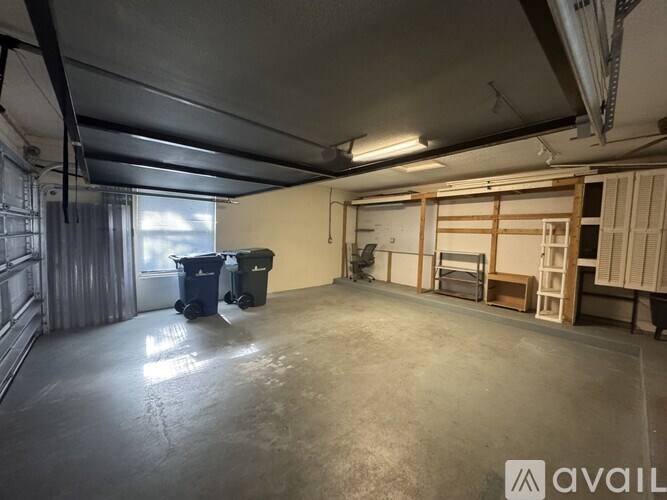 A large, empty room with a concrete floor and walls, featuring a window with curtains, two trash bins, and a ladder.
