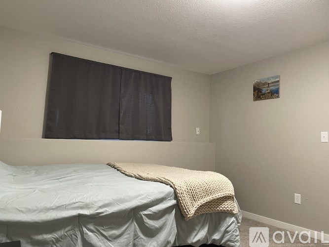 A bedroom with a bed covered in a blue sheet and a blanket, a window with black curtains, and a small picture on the wall.
