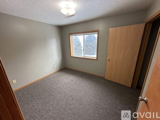 A room with a carpeted floor, a window with a view of trees outside, and a door on the right side.