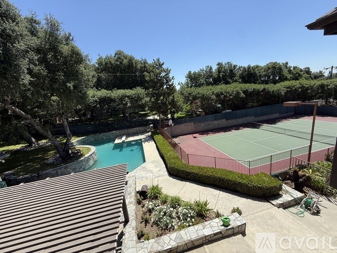 A tennis court is surrounded by a pool and greenery.