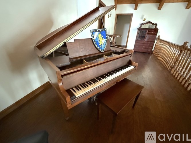 A grand piano with a shield on top is situated in a room with wooden flooring and a wooden railing.