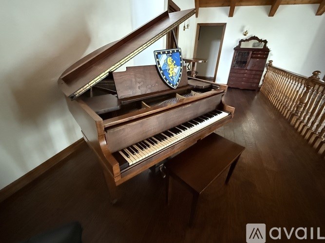 A grand piano with a shield on top is situated in a room with wooden flooring and a wooden railing.