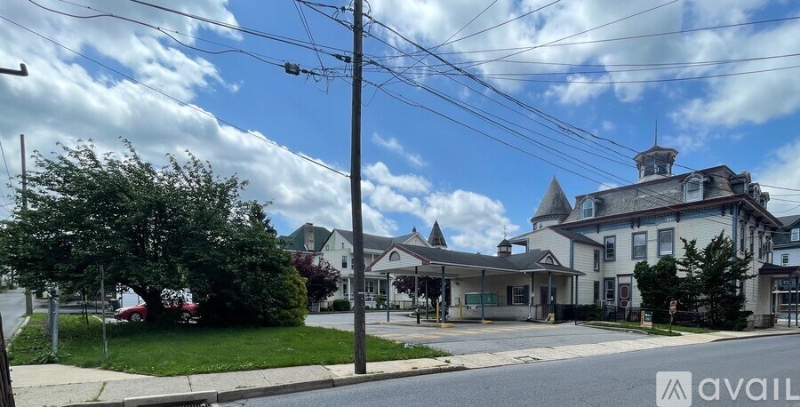 A large white building with a tower-like structure on the roof is surrounded by trees and a street in front.