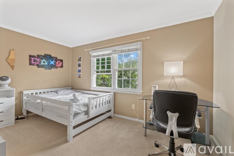 A bedroom with a white bed, a desk with a computer chair, and a window with a view of trees.