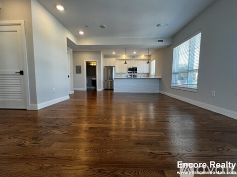 A spacious room with wooden flooring and a kitchen area in the background.