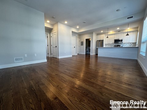 A spacious room with wooden flooring and a kitchen area in the background.