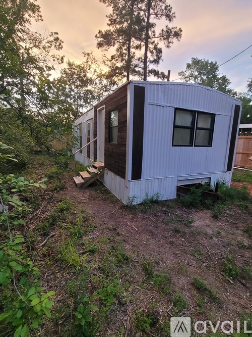 A small white house with a brown door and windows is surrounded by vegetation.