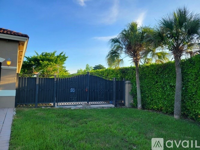 A black fence with a gate is surrounded by greenery and palm trees.
