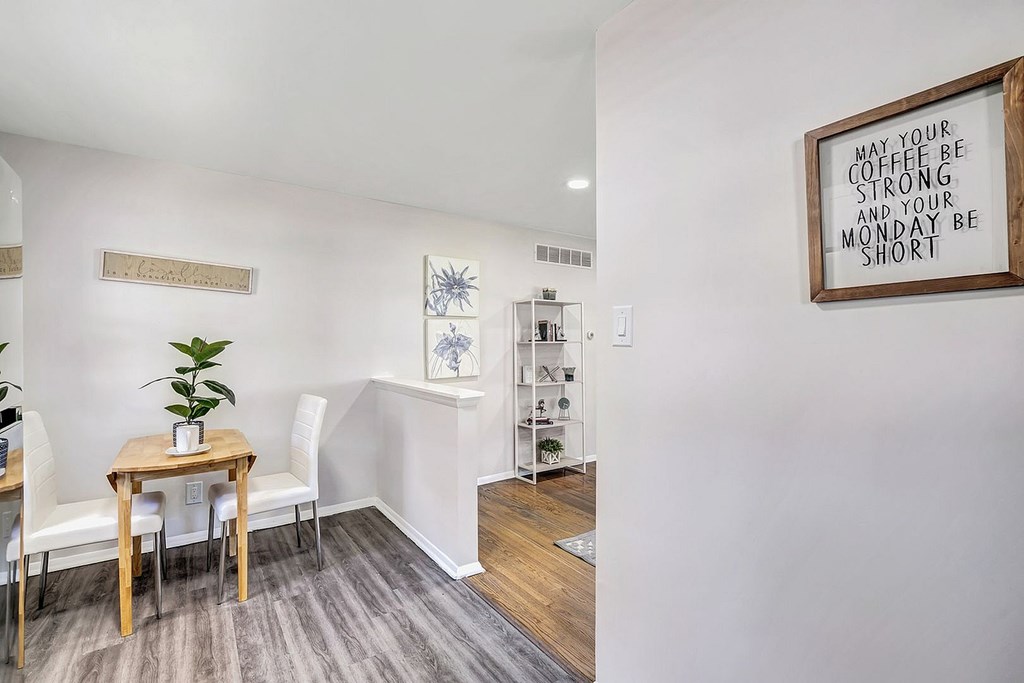 A dining room with a wooden table and chairs, a framed sign on the wall, and a potted plant on the table.