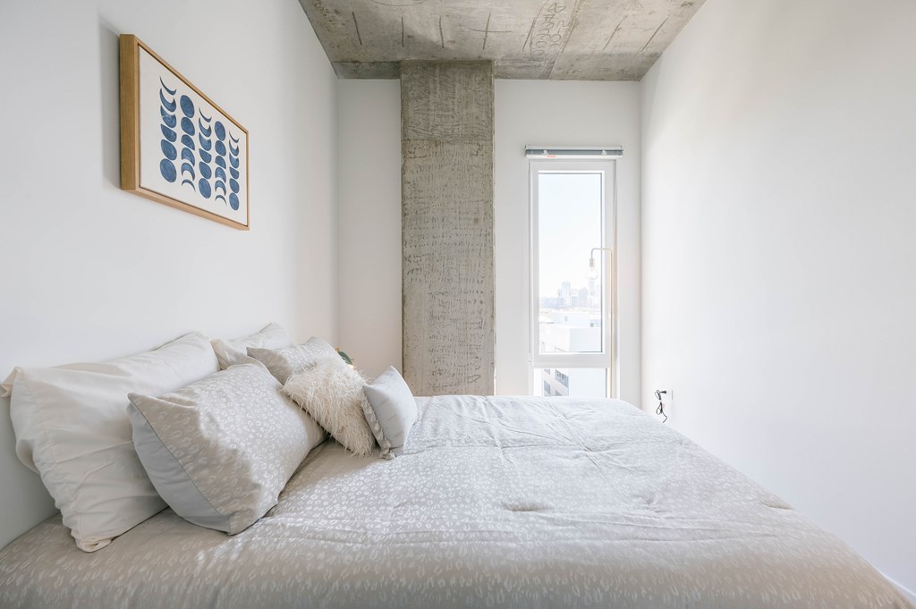 A bed with a white comforter and pillows in a room with a framed picture on the wall.