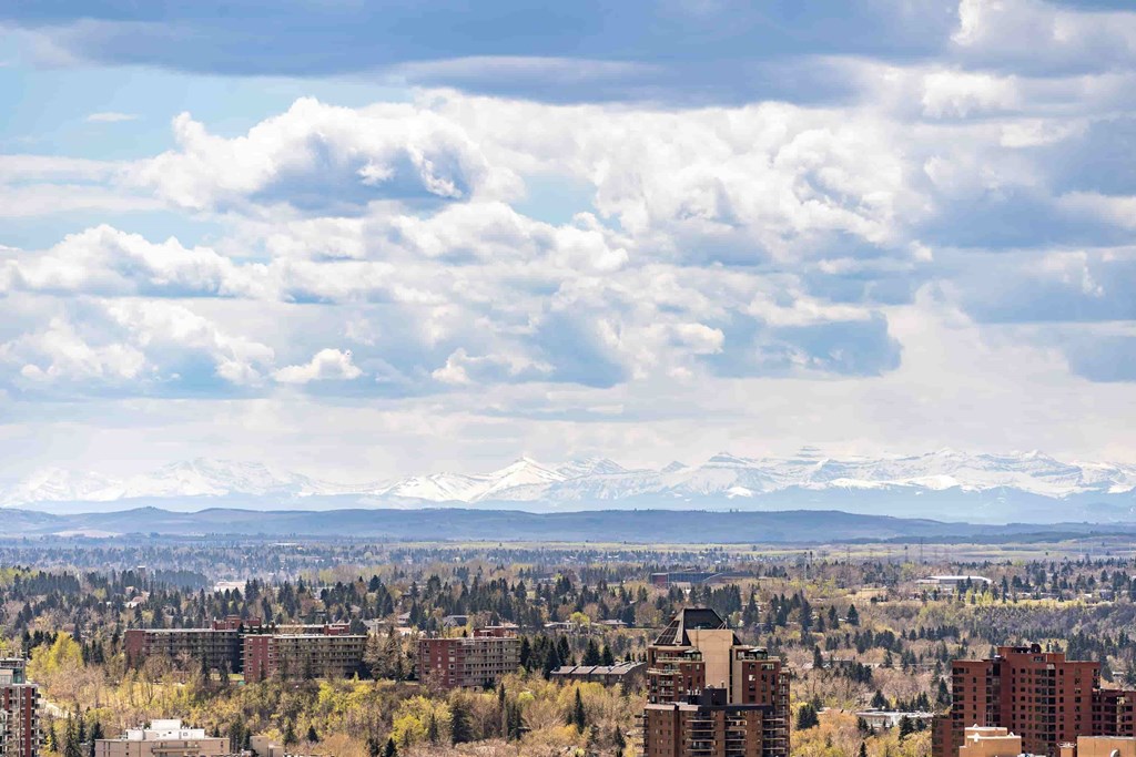 A cityscape with buildings and trees in the foreground and snow-capped mountains in the distance under a cloudy sky.