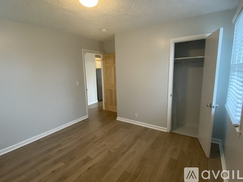 A room with wooden floors and white walls, with a door on the right and a window with blinds on the far right.