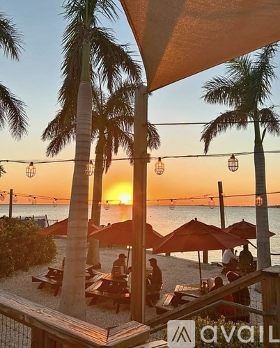 A beach scene with palm trees and people sitting at tables under umbrellas.