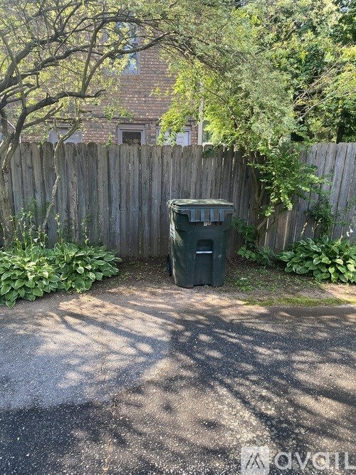 A green trash bin sits in a backyard next to a wooden fence.
