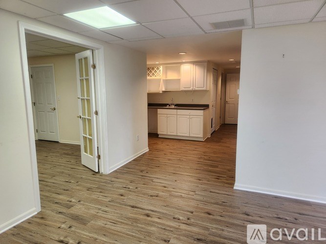 A room with wooden flooring and white walls, with a kitchen area visible in the background.