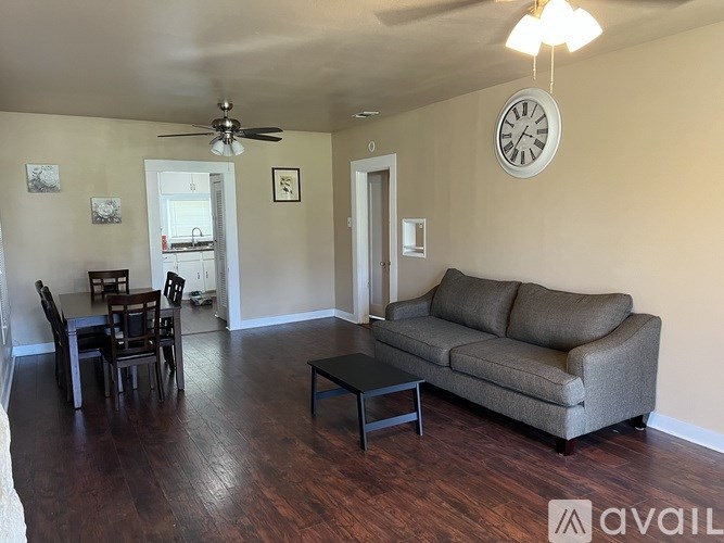 A living room with a grey couch, a dining table with chairs, and a clock on the wall.