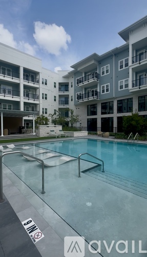 A large swimming pool is surrounded by palm trees and lounge chairs in front of a multi-story apartment building.