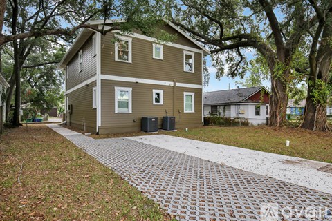 A two-story house with a brown and beige exterior is surrounded by trees and has a brick driveway.