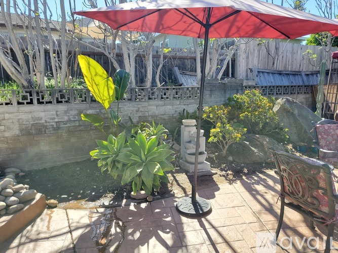 A red umbrella provides shade over a potted plant and a bench in a sunny outdoor setting.