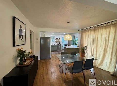 A living room with a glass table and chairs, a framed picture on the wall, and a dining area in the background.