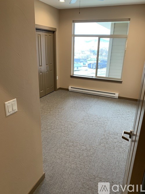 A room with a carpeted floor, a window with a view of buildings outside, and a door on the left side.