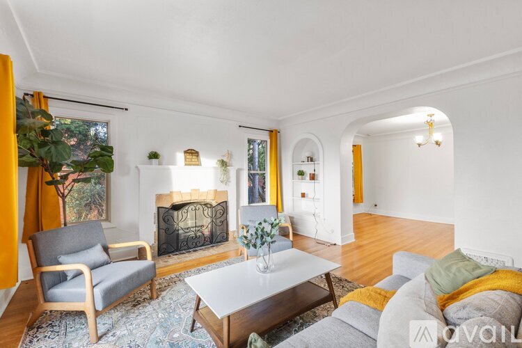 A living room with a grey sofa, a grey armchair, a white coffee table, a fireplace, and a large window with yellow curtains.