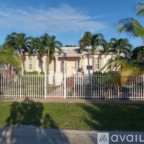 A white house with a fence in front of it and palm trees in the background.