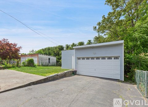 A large garage with a white door is situated in a driveway with a green fence and trees in the background.
