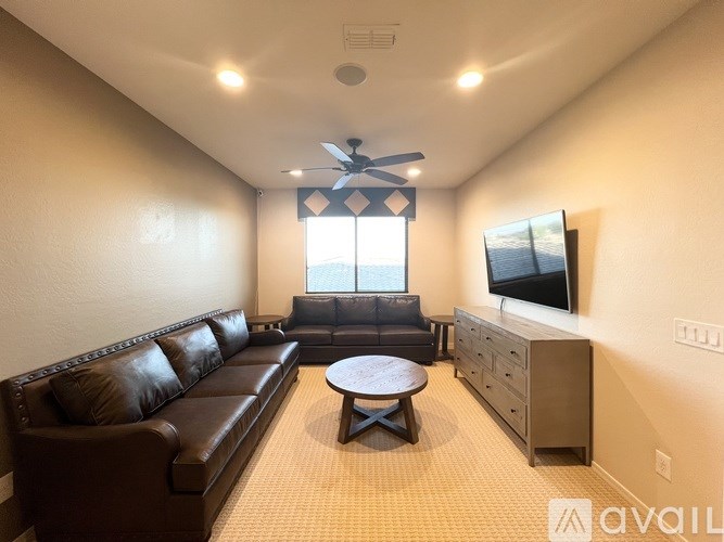 A living room with a brown leather couch, a black ottoman, a TV, and a ceiling fan.
