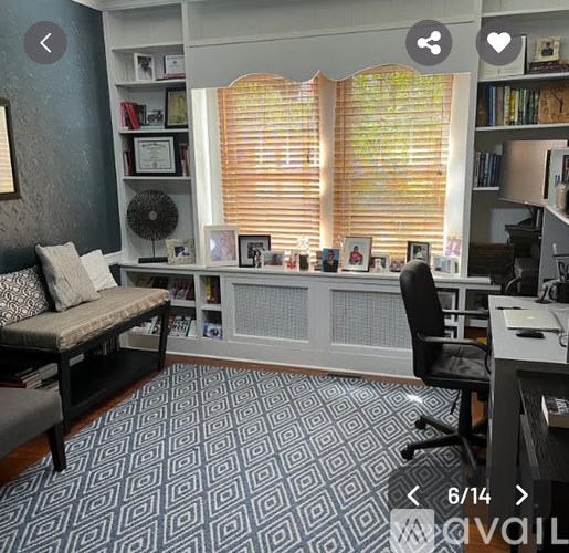 A room with a patterned carpet, a window with blinds, a desk with a chair, and a bookshelf with books.