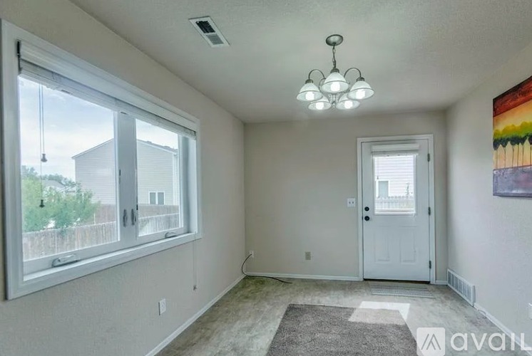 A room with a white door, a window with a view of a fence and trees, a chandelier, and a painting on the wall.