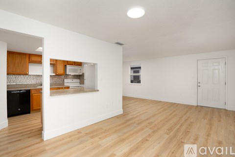 A room with wooden flooring and white walls, leading to a kitchen area.